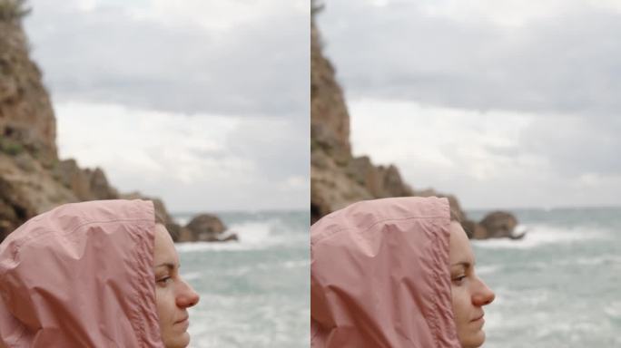 垂直视频。一位身穿粉色外套的年轻女子凝视远方，陷入沉思。当她坐在岸边时，大浪冲击着岩石。缓慢的运动。