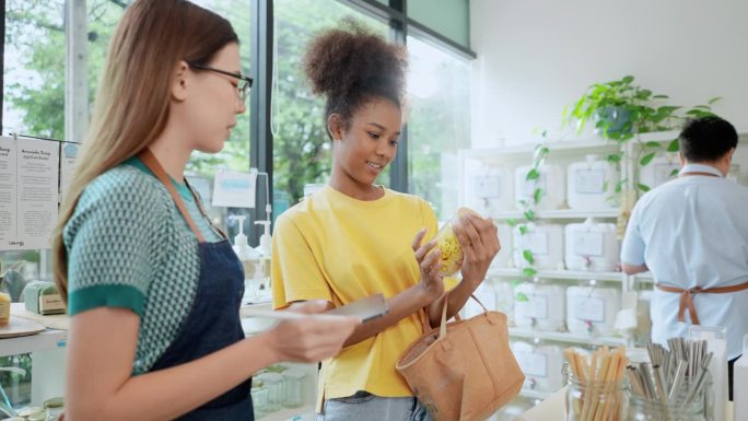 亚洲女销售代表向穿着黄色t恤的非洲女顾客推荐小罐子里的零食，而一名不认识的男子在补充店检查其他产品的