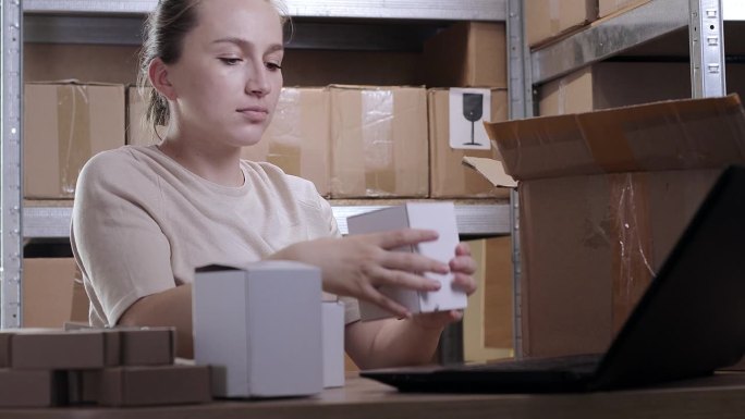一个女人在仓库里打开一个硬纸板箱，看了看货物，列了一份清单。跟单员叙述箱子里的物品