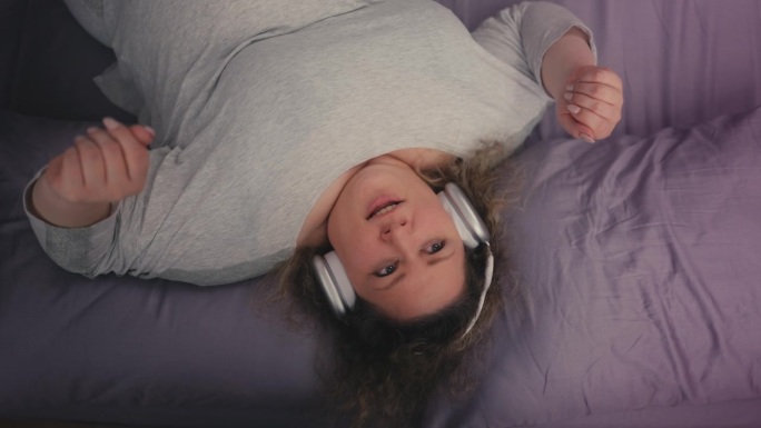 俯视图快乐超重女人听音乐和唱歌在床上的耳机