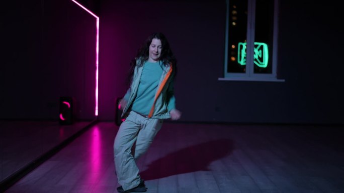 住相机。一个欢快的女孩在黑暗的舞蹈大厅里用霓虹灯照亮的镜子跳着充满活力的嘻哈舞。