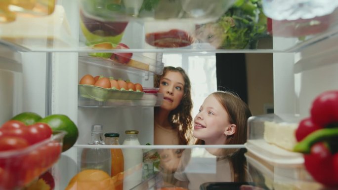 妇女和女孩用健康食品打开冰箱门并服用产品。快乐家庭做饭和选择冰箱食材。微笑的人们选择储存食物的生活