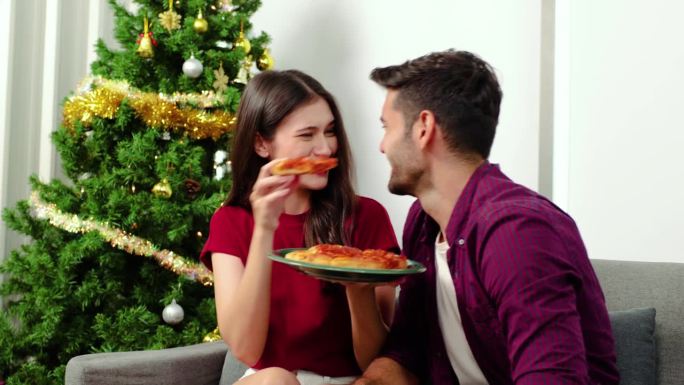 圣诞快乐，新年快乐，年轻夫妇在家里吃晚饭。两个白种人在家里的圣诞树旁喝啤酒，吃披萨。庆祝节日和团聚。