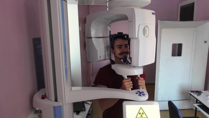 在牙科诊所，一名年轻的白人男子微笑着坐在计算机头部断层扫描前