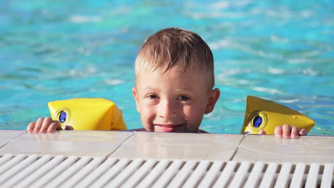戴着臂章在游泳池里洗澡和游泳的白人男婴的脸部特写。孩子趴在泳池边看镜头。暑假的休闲活动。享受快乐的童