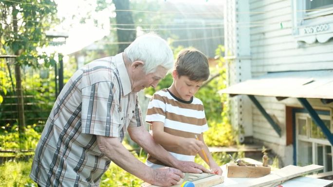 一位白发苍苍的老人在户外教一个男孩做木工活。爷爷教孙子做木匠，用尺子量木板，用铅笔划线。