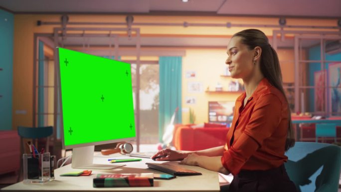 高加索女创作者使用桌面电脑与绿色色度键屏幕在她的彩色快乐的公寓。自由职业女性致力于她的项目，她对进展