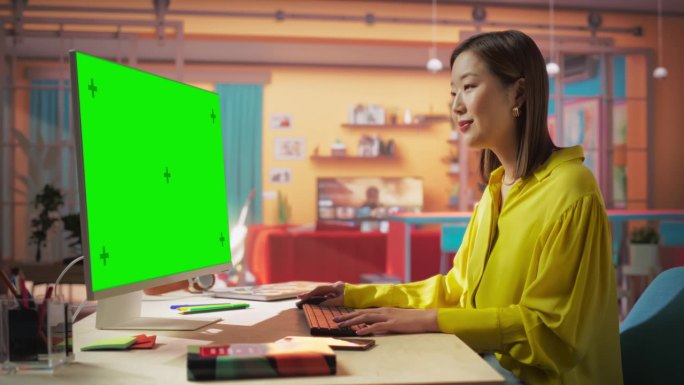 富有创造力的年轻亚洲女性坐在她的办公桌前，使用带有绿屏模型的台式电脑。在彩色公寓里用彩色键显示电脑工