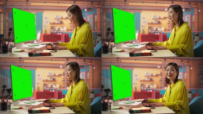 富有创造力的年轻亚洲女性坐在她的办公桌前，使用带有绿屏模型的台式电脑。在彩色公寓里用彩色键显示电脑工