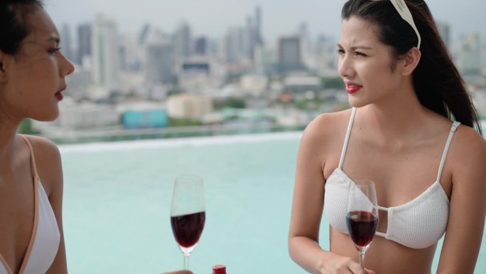 年轻的成年美女们穿着比基尼坐在泳池边，喝着红酒聊天，在一个豪华的度假胜地放松，庆祝他们一生中的成就。