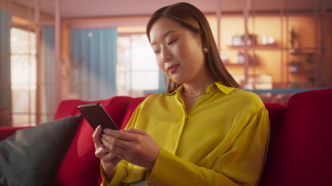 年轻美丽的亚洲女性在客厅的肖像，休息和使用智能手机。创意女孩查看社交媒体，与朋友聊天，上网浏览有趣的