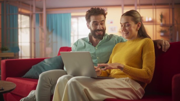 年轻夫妇坐在家里的沙发上使用笔记本电脑的肖像。订婚夫妇在网上为他们的婚礼准备购物，讨论使用技术的不同