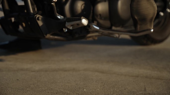 一个男人的腿伸到了摩托车的脚踏板上。自行车细节特写。黑色摩托车在温暖的阳光下。摩托车在柏油路上。车辆