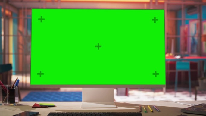 现代桌面电脑显示器与绿色屏幕模拟显示站在桌子上与键盘，鼠标，笔，笔记本和耳机。舒适的家庭办公室在色彩