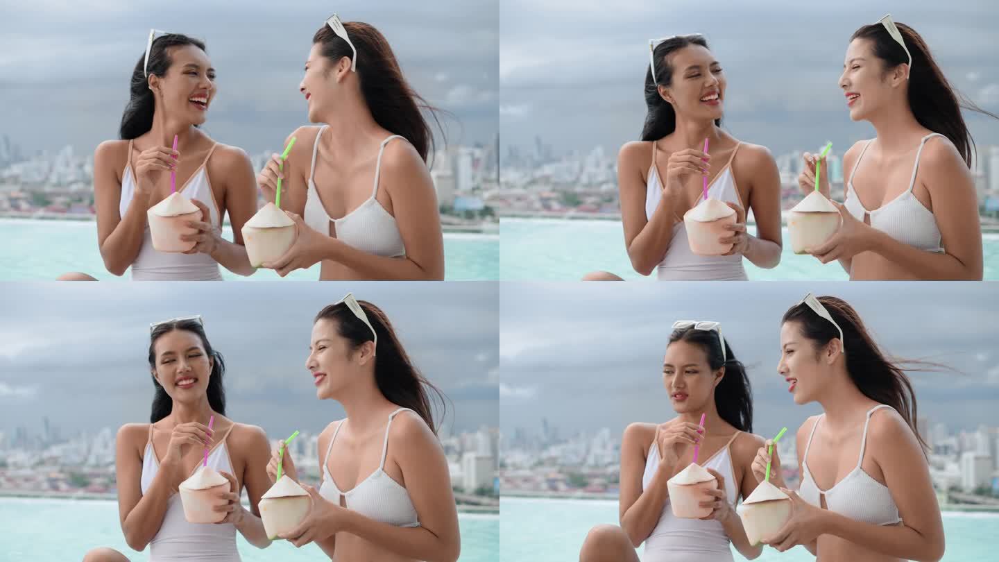 两个美丽的女性朋友穿着白色的泳衣，在屋顶的游泳池边喝着椰子汁，以城市为背景，幸福地谈论着过去一年的生