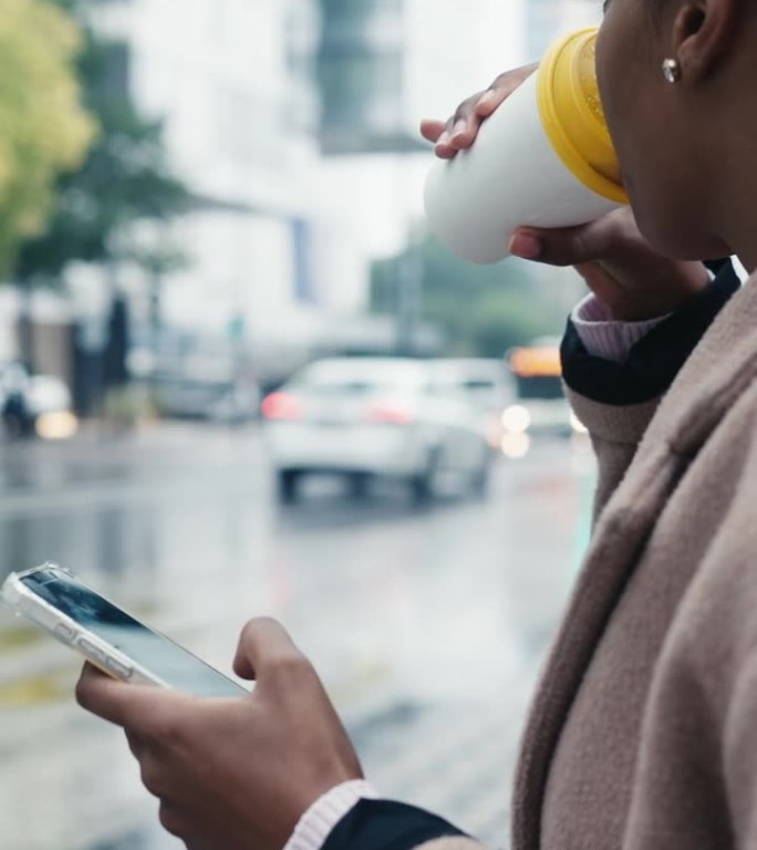 雨季里，穿着长外套的年轻女子坐在公交车站边喝咖啡边玩手机