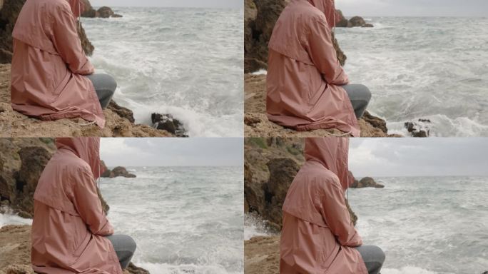 一个孤独的女人，穿着粉红色的夹克，戴着兜帽，坐在一块岩石上，凝视着汹涌的大海。