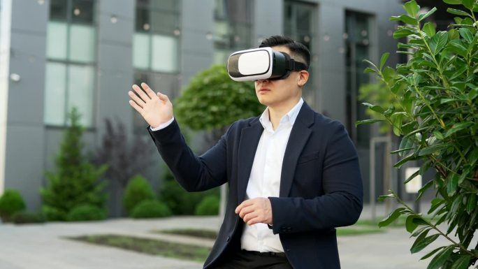一位亚洲商人坐在办公大楼附近街道的长椅上，戴着虚拟现实眼镜在虚拟现实模拟器中工作