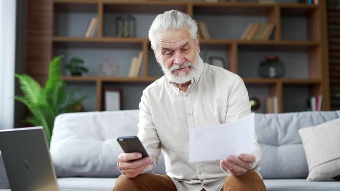 快乐的老人坐在家里的房间里用智能手机使用手机应用程序支付账单