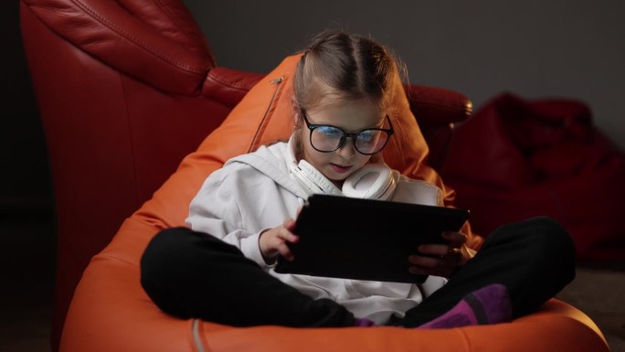 兴奋的少女坐在家里的沙发上，用数字平板电脑技术设备玩游戏。情绪化的孩子戴着眼镜拿着pad电脑上网。儿