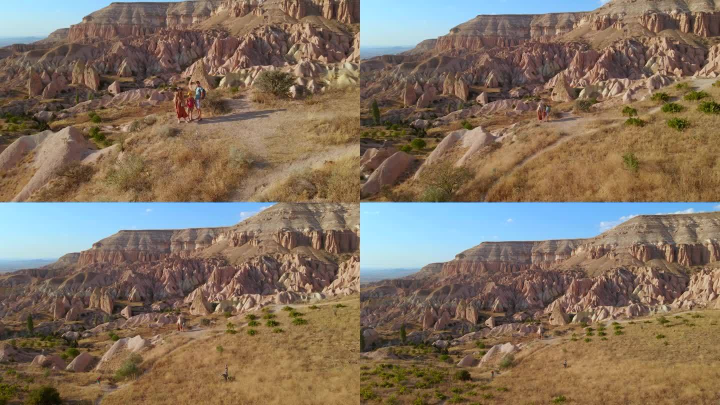 一个游客家庭徒步穿越卡帕多西亚戈雷梅附近的红色或粉红色山谷。以暖色调绘制的景观展示了独特的地质构造和