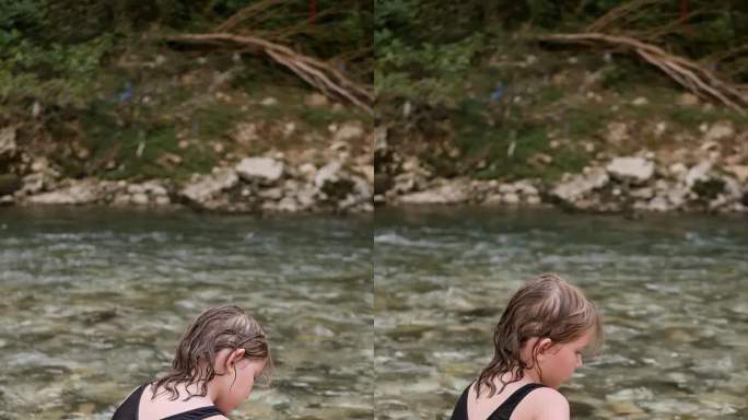 垂直视频显示，一个八岁的小女孩穿着连体泳衣，坐在山河的岩石岸边，水流湍急，她的脚浸在冰冷的水中。