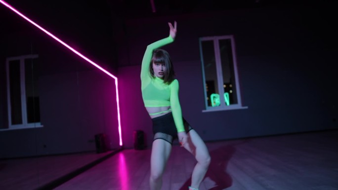 住相机。一个年轻的女孩穿着明亮的舞台服装，在霓虹灯照亮的黑暗舞蹈大厅里跳电臀舞。