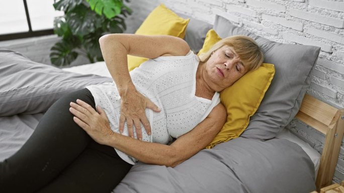 一个中年金发女人，肚子疼得厉害，躺在床上不高兴地摸着肚子，说明了室内疾病的挣扎。