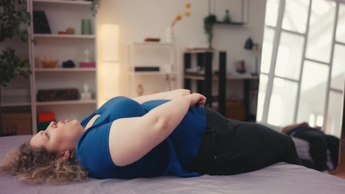 滑稽超重的女人躺在床上，试图穿上紧身牛仔裤