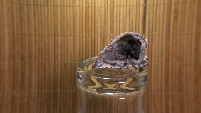 紫色晶洞晶体在一个翻转的玻璃杯上缓慢旋转，在一个模糊的棕色烟叶背景