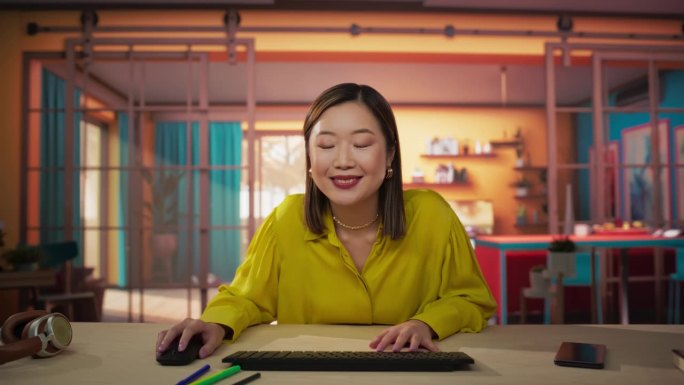 屏幕视角:美丽的亚洲女人坐在一张桌子上，在一个舒适的彩色公寓和使用电脑。微笑的女性成年人点头，在网上