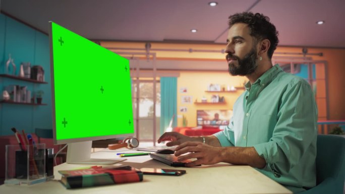 年轻英俊的男子从家里工作的桌面电脑与绿屏模拟显示。微笑的男性查看公司账户，给同事发信息，做创意项目