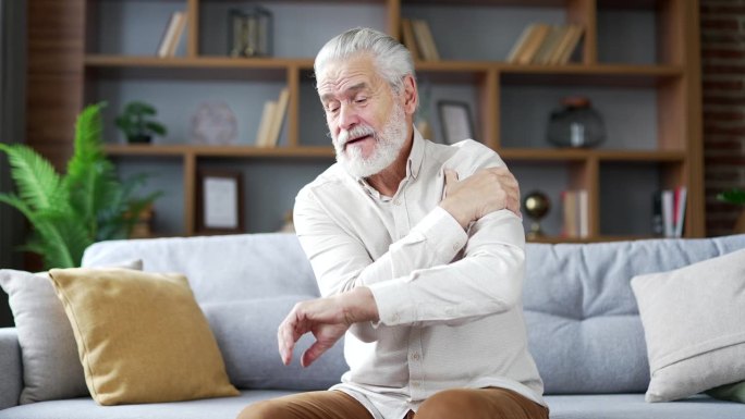 一位白发老人坐在家里客厅的沙发上，肩膀剧痛。