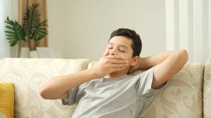 一个穿着灰色t恤又累又伤心的少年坐在沙发上看电视。无聊的少年双手抱头打哈欠。
