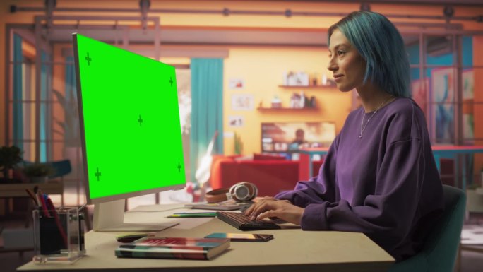 静态肖像美丽时尚的女人与蓝头发使用现代桌面电脑与绿屏模拟显示。创意女性设计师和程序员从时尚之家工作