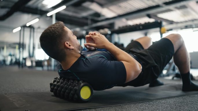 肌肉发达的男运动员穿着运动服在健身房做泡沫滚轮练习以缓解背部疼痛