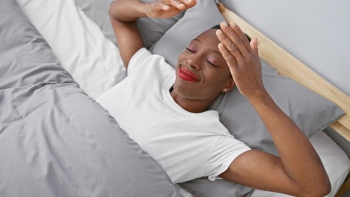一个非裔美国妇女躺在床上，双手蒙住眼睛，笑得欢快而有趣。盲目的概念。在卧室