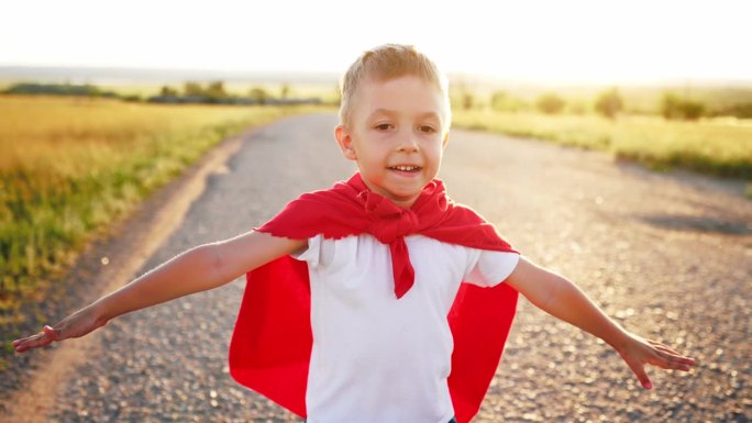 男孩扮演超级英雄。快乐的小男孩在玩超级英雄。日落时分，小孩穿着红斗篷跑过马路。在大自然中穿着红色斗篷