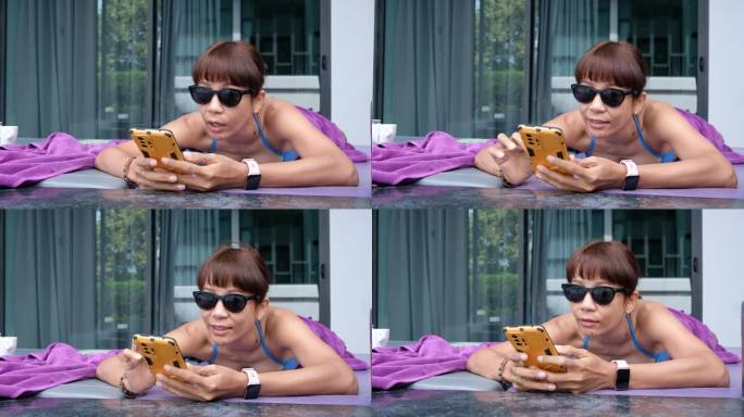 穿着泳装、戴着墨镜的亚洲女性躺在酒店阳台的靠垫上玩智能手机