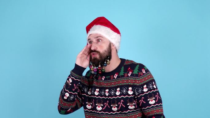 快乐的年轻胡子男人戴着圣诞老人的帽子摆姿势，试图听到你无意中听到孤立的蓝色背景。