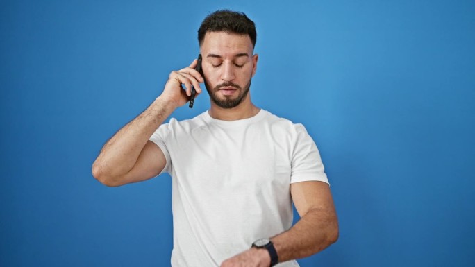 年轻的阿拉伯男子正在用智能手机通话，看着孤立的蓝色背景手表