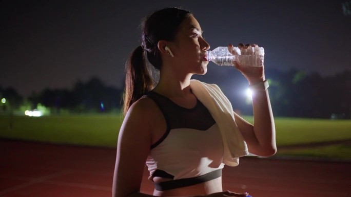 年轻的亚洲女性在跑道上跑了一晚后，停下来喝水，查看智能手表上的跑步数据