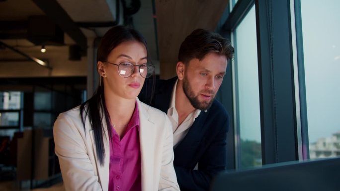 一个身穿白色夹克和粉色衬衫的黑发女孩，戴着圆框眼镜，和一个穿蓝色夹克的男人坐在一起，在办公室的全景窗