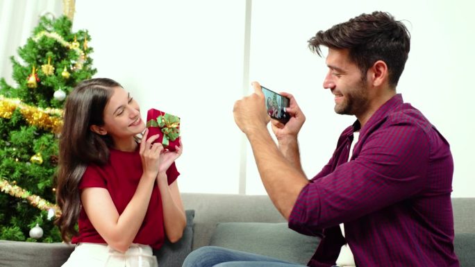 年轻的白人男友坐在圣诞树旁，用智能手机拍下女友拿着礼物的照片。新年和圣诞节的美好回忆，快乐的相聚时光