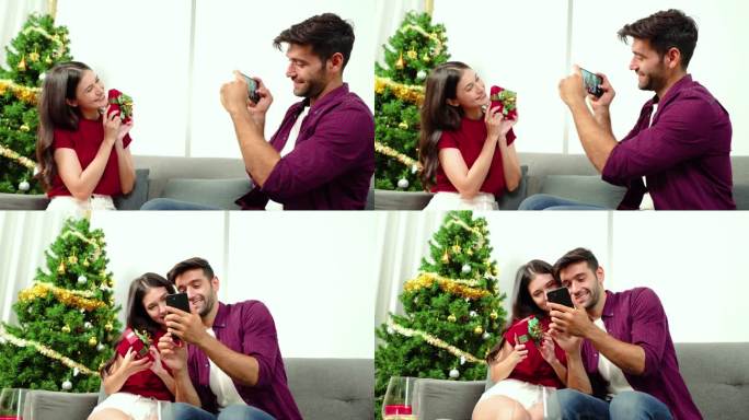 年轻的白人男友坐在圣诞树旁，用智能手机拍下女友拿着礼物的照片。新年和圣诞节的美好回忆，快乐的相聚时光