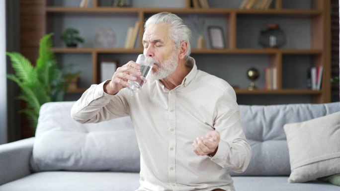 生病的白发大胡子老人坐在家里客厅的沙发上，一边吃着止痛药，一边用杯子喝水。