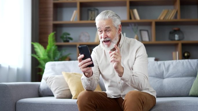 一位头发花白的老人坐在家里客厅的沙发上，用智能手机收到了好消息。
