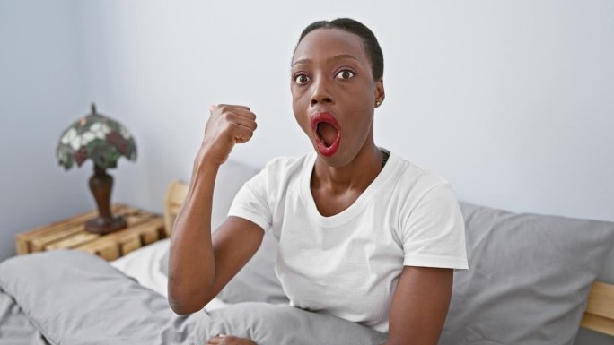 惊讶的非裔美国女人在卧室里，躺在床上带着惊讶的表情，眩晕地指着，嘴巴张得很大!