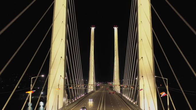 一座斜拉桥，有高大的混凝土塔，夜间有建筑照明。天桥上架桥塔之间的路基上，汽车来往于下面