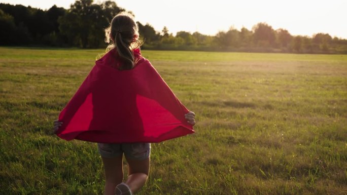 女孩扮演超级英雄。快乐的女孩在玩超级英雄。日落时分，孩子们穿着红斗篷跑过绿色的田野。在大自然中穿着红